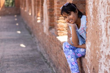 Mavi elbiseli Hintli güzel bir kız çocuğu gülen yüzüyle tuğla duvardan dikizliyor.