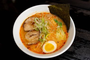 Bu resimde baharatlı Tonkotsu ramen kasesi var. İçinde domuz eti, haşlanmış yumurta, yosun, yeşil soğan ve diğer malzemeler var. Beyaz bir kasede servis ediliyor..