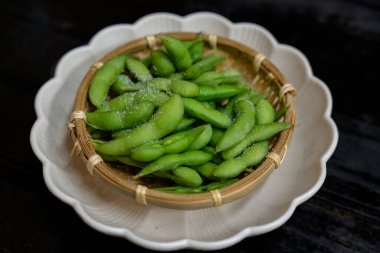 Bu resim, dekoratif beyaz bir tabağa yerleştirilmiş deniz tuzlu yeşil edamame fasulyeleriyle dolu küçük bir sepeti gösteriyor. Sunum basit ama iştah açıcı, edamamanın canlı yeşil rengini vurguluyor.