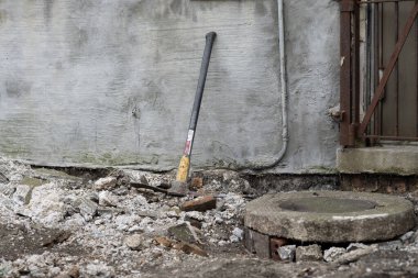 Bir inşaat çekici molozlarla çevrili gri beton bir duvara yaslanır. Sahne, hammaddelerin etrafa saçıldığı devam eden bir yıkım veya yenileme projesine işaret ediyor..