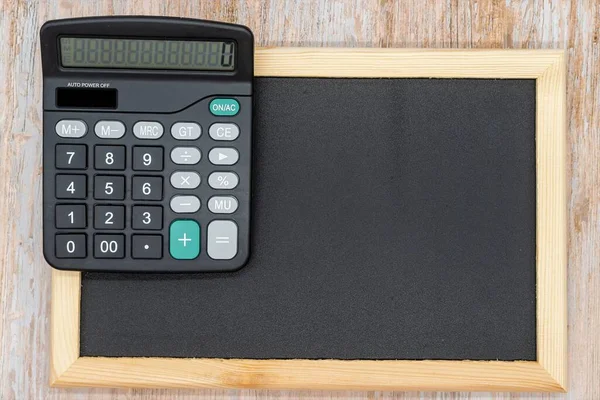 Black wooden blackboard and electronic calculator. Top view. Copy space. Taxes, mathematics, investments, costs, payments.