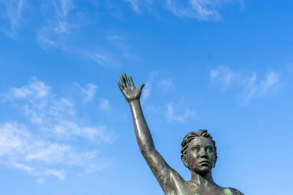 Bronze statue of a child tightrope walker in a blue sky with clouds. Detail of the hand upwards