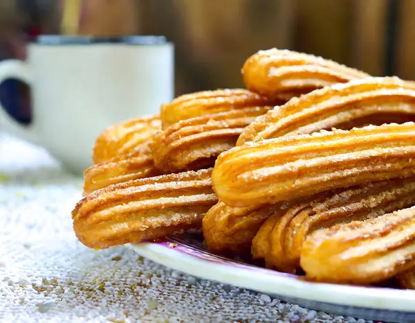 Tepesine şeker serpiştirilmiş bir tabak churros bir bardağın yanında duruyor.