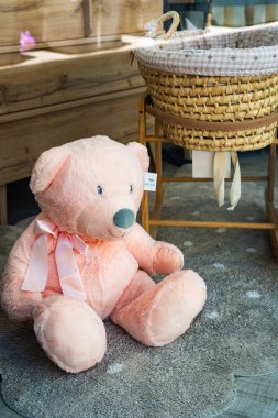 Pembe bir oyuncak ayı, bir çocuk odasında yerde, hasır bir beşiğin yanında, sıcaklığı ve şefkati çağrıştırıyor.