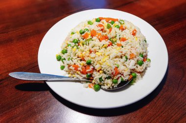Boiled white rice mixed with colorful vegetables, onion, carrot, green peas, corn, green beans in a white plate on the table