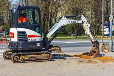Bobcat mini excavator with bulldozer, rubber tracks dig sand on sidewalk in city. Panevezys. Lithuania. 19. 04. 2023. clipart