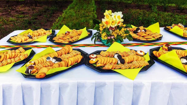 Trays with various cookies, cupcakes and cakes outdoors on a table with a white tablecloth and a bouquet of daffodils at a celebration