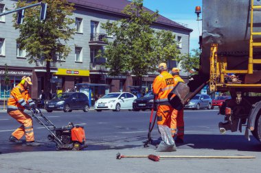 Turunculu üç yol inşaat işçisi, bir yol silindiri ile bir şehir caddesinde asfalt tamir ediyorlar. Arabalar binalar ve ağaçlar yollarda ilerlerken geçiyor. Panevezy 'ler. Litvanya. 02 numara. 06 numara. 2023.