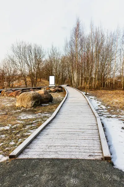 A wooden path leads through the snow-covered park with snow-covered trees and bushes. An empty signpost stands at the end of a dense, bare park path. The footpath is covered with thick snow.