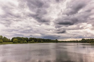 Göl, ağaçlar ve bulutlu gökyüzü olan sakin bir manzara. Durgun su bulutlu gökyüzünü yansıtırken sahne sükunet yayar. Hışırdayan yapraklar ve ağaç türlerinin karışımı doğanın sakin güzelliğini artırır..
