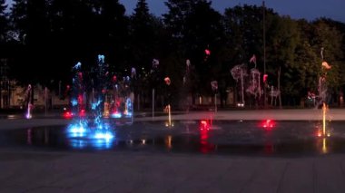 Canlı renkli fıskiyeler gece şehir meydanını aydınlatıyor, büyüleyici bir yavaş çekim görüntüsü sergiliyor..