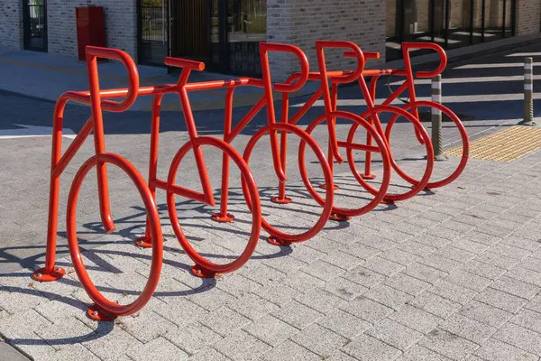 Altı sıra boş kırmızı metal bisiklet park yeri, bisiklet şeklinde tasarlanmış, kırsal tuğla bir binaya karşı dizilmiş, kentsel altyapı ve modern tasarım sergileniyor..