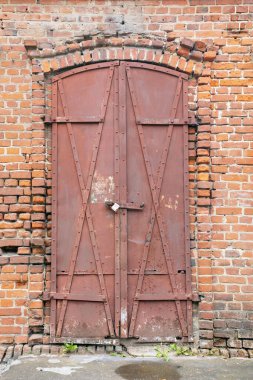 Eski kırmızı ağır metal kapı iki büyük çapraz metal plakayla süslenmiştir. Yüzeyi paslanmış ve soyulmuş boya izleri gösteriyor, yıpranmış tuğla bir duvara karşı dizilmiş, bir tarih algısı uyandırıyor..