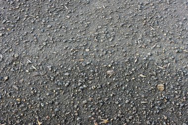 Asfalt dokusuna yakın çekim. Yüzeyin her tarafına dağılmış küçük çakıl taşları ve kayalar doğal ve engebeli bir görünüm yaratıyor..