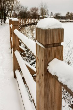 Taze karla kaplı ahşap bir çitle kaplı bir patika donmuş bir göle götürür. Sessiz kış ortamı, gri gökyüzünün altında yapraksız ağaçlarla çevrilidir..