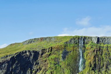 Şelale, parlak yeşil yosun ve kayalık araziyle çevrili sarp kayalıkların üzerinden zarif bir şekilde akar. Açık mavi gökyüzü İzlanda 'daki bu doğa harikası için mükemmel bir zemin..