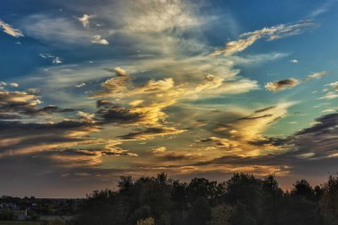 The sky is painted with vibrant colors as the sun sets, casting golden hues among the clouds. Dark silhouettes of trees mark the horizon, creating a peaceful atmosphere.