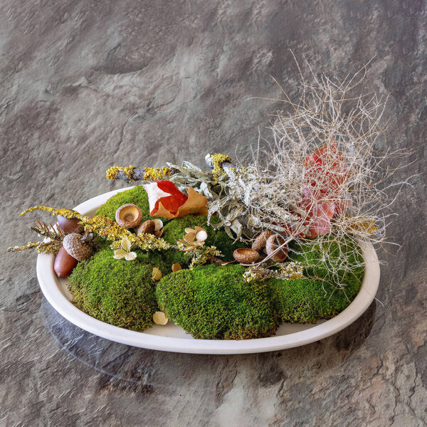 The autumn composition showcases lush green moss, a variety of oak acorns, and delicate botanicals, carefully arranged on a marble surface. This natural centerpiece highlights seasonal beauty.