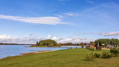 Bereketli bir göl kenarı, verimli yeşil çimenler, berrak mavi su ve sonbaharı yansıtan renkli ağaçlara sahiptir. Gökyüzü ince bulutlarla doludur. Kupiski. Litvanya. 07.09.2024.