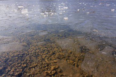 Buz parçaları donmuş bir gölün yüzeyinde nazikçe yüzerler. Aşağıda, sakin ve buzlu sualtı manzarası görülebilir, kışın doğal güzelliğini gözler önüne seriyor..