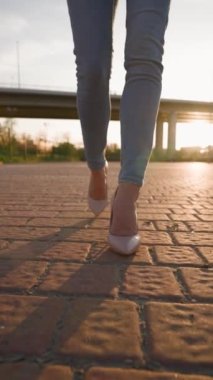 Dikey Video, Yüksek Topuklu Ayakkabılı İnce Kadın Bacakları Şehir Parkı 'nda Günbatımı Yürüyüşü. Gün batımında şehrin caddesinde yürüyen güzel bacaklı kadın. Tanınmayan kişi. Yavaş Hareket.