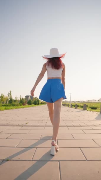 垂直视频 后视镜美丽的瘦女人走在一个城市公园与优雅的步态 穿着蓝色短裙的有诱惑力的女孩自信地走在夕阳西下的城市大街上 慢动作 — 图库视频影像