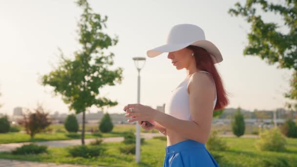 快乐女人与瘦身材微笑和步行在城市公园使用手机社交网络 穿着白帽散步的漂亮姑娘在夏城周围用智能手机 慢动作 — 图库视频影像