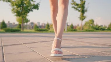 Yüksek topuklu sandaletlerle baştan çıkarıcı kadın bacakları güneşli şehir parkında zarafetle yürüyor. Sexy Slim Woman Walks with a Confident Gait Along a City Street. Yavaş Hareket.