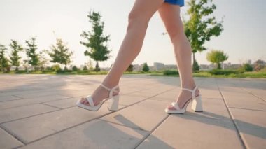 Yüksek topuklu sandaletlerle baştan çıkarıcı kadın bacakları güneşli şehir parkında zarafetle yürüyor. Sexy Slim Woman Walks with a Confident Gait Along a City Street. Yavaş Hareket.