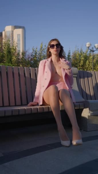 在阳光明媚的日子里 坐在城市公园的长椅上 穿着粉红工作服的漂亮女人坐在长椅上 时尚与美感概念 垂直录像 慢动作 — 图库视频影像