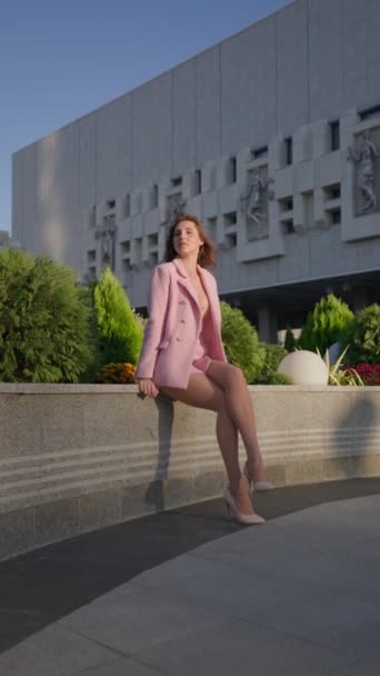 在阳光明媚的日子里 坐在城市公园的栏杆上 身穿粉红工作服的漂亮女人坐在那里 时尚与美感概念 垂直录像 慢动作 — 图库视频影像