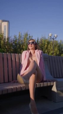 Güneşli bir günde bir şehir parkının bankında otururken Pembe İş Kıyafeti Pozisyonunda Güzel Kadın. Moda ve Güzellik Konsepti. Dikey Video, Yavaş Hareket.