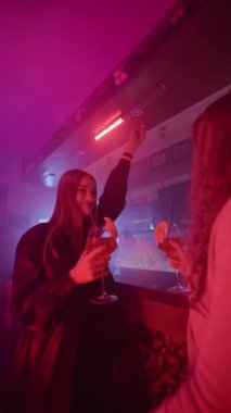 Dikey Video, İki Kız Arkadaş Bir Neon Lighting Gece Kulübü 'nde Ellerinde Barın Yanında Kokteyllerle Dans Ediyor. Gece Barındaki Partide Eğlenen Mutlu Kadınlar. Yavaş Hareket.