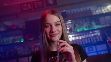 Neon Lighting 'deki bir barda Kırmızı Kokteyl İçen Çekici Gülümseyen Kız portresi. Arkaplanda Alevler ve Yangın. Gece Rahatlama ve Eğlence Konsepti. Yavaş Hareket.