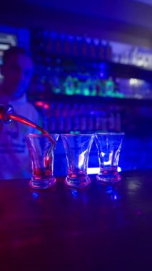Dikey Video, Guy Barmen, Blue Neon Lighting 'deki bir barın üzerinde üç şeffaf bardağa alkollü kokteyl döküyor. Bir gece kulübünde eğlence ve eğlence kavramı. Yavaş Hareket.