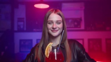 Bardaki tezgahta oturan sevimli mutlu sarışın kız. Kırmızı Alkollü Kokteyl içip gülümsüyor. Neon Lighting 'de bir gece barı. Yavaş Hareket