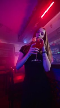 Güzel Mutlu Genç Kız Gece Kulübü 'nde Gülümseyip Dans Ediyor ve Kırmızı Alkolik Kokteyli İçiyor. Arka planda Neon Lighting Bar Sayacı var. Yavaş Hareket, Dikey Video
