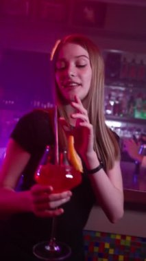 Güzel genç kız elinde kırmızı bir kokteyl ile gece kulübünde dans ediyor. Arka planda Bar Sayacı ve Barmen Ateş Kullanarak Alkollü İçecekleri Hazırlıyor. Yavaş Hareket, Dikey Video