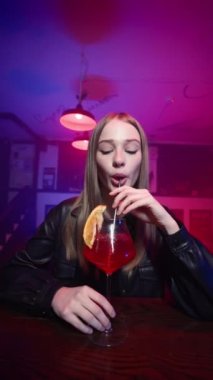 Bardaki tezgahta oturan sevimli mutlu sarışın kız. Kırmızı Alkollü Kokteyl içip gülümsüyor. Neon Lighting 'de bir gece barı. Yavaş Hareket, Dikey Video.