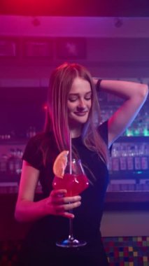 Gece kulübünde elinde kırmızı kokteylle gülümseyen ve dans eden güzel genç kız. Arka planda bar tezgahı var. Yavaş Hareket, Dikey Video