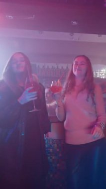 Dikey Video, İki Kız Arkadaş Bir Neon Lighting Gece Kulübü 'nde Ellerinde Barın Yanında Kokteyllerle Dans Ediyor. Gece Barındaki Partide Eğlenen Mutlu Kadınlar. Yavaş Hareket.