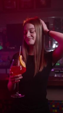 Dikey Video, Neon Lighting 'deki Barda Kırmızı Kokteyl İçen Çekici Gülümseyen Kız. Arkaplanda Alevler ve Yangın. Gece Rahatlama ve Eğlence Konsepti. Yavaş Hareket.