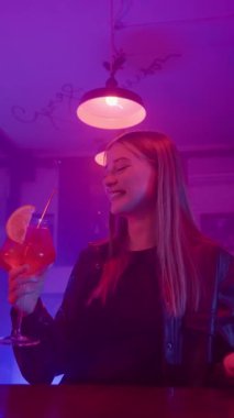 Bardaki tezgahta oturan sevimli mutlu sarışın kız. Kırmızı Alkollü Kokteyl içip gülümsüyor. Neon Lighting 'de bir gece barı. Yavaş Hareket, Dikey Video.