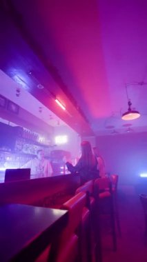 İki Yetişkin Kadın Gece Barında Bir Barmenle Konuşuyor. Mor ve Mavi Neon Işıklandırma. Boş zaman ve alkol konsepti. Dikey video. Yavaş Hareket.