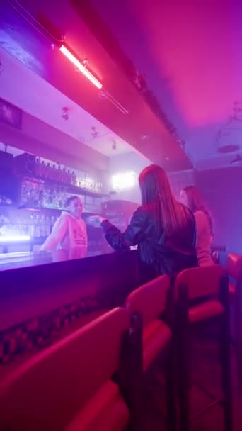 2人の若い大人の女性がナイトバーでバーテンダーと話す 紫と青のネオン照明 レジャー アルコール飲料コンセプト 垂直ビデオ スローモーション — ストック動画