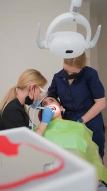 Genç Yetişkin Kadın Bir Diş Kliniğinde Bir Kadın Diş Hekimi ve Asistanı tarafından Temizlik Prosedürü Yaptırıyor. Ortodontist ve Tıp Konsepti. Diş tedavisi için. Dikey Video, Yavaş Hareket