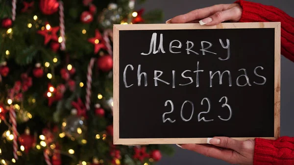 Marry Christmas banner concept. Female hands in red sweater hold chalk board with text Merry Christmas 2023 on background of beautiful festive Christmas tree decorated with red toys and garlands.