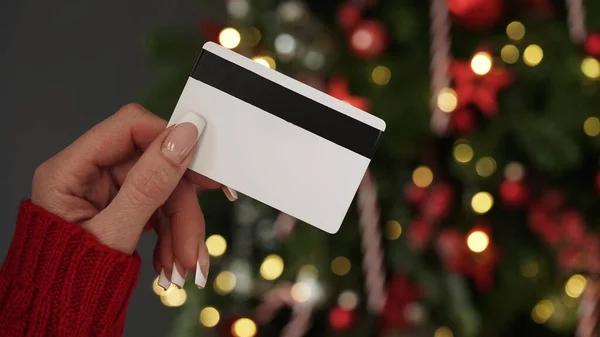 Hands of a woman in a red sweater with a credit card on the background of a Christmas tree close-up. Christmas and New Year shopping online, credit card payment. Buying gifts online