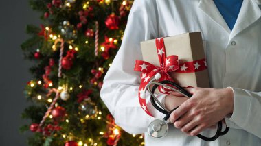 Noel ve yeni yıl tıbbi afişi. Beyaz önlüklü bayan doktor, steteskop elinde kırmızı fiyonkla arka planda güzel dekore edilmiş Noel ağacını tutuyor. Kopya alanı.