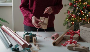 Burgonya rengi süveterli bir kadın, içinde hediyeler olan bir sürü kutuyu güzel kağıt ve kurdelelerle paketliyor, bir Noel ağacının arka planında fiyonklar. Noel, yeni yıl tatili, hediye paketi..